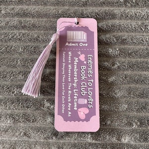 May include: A pink and purple bookmark with a tassel. The bookmark has a ticket design and says "Enemies to Lovers Book Club Membership: Lifetime" and "Where: Wherever The Rivals Are At" and "Forever Denying Their Love For Each Other".