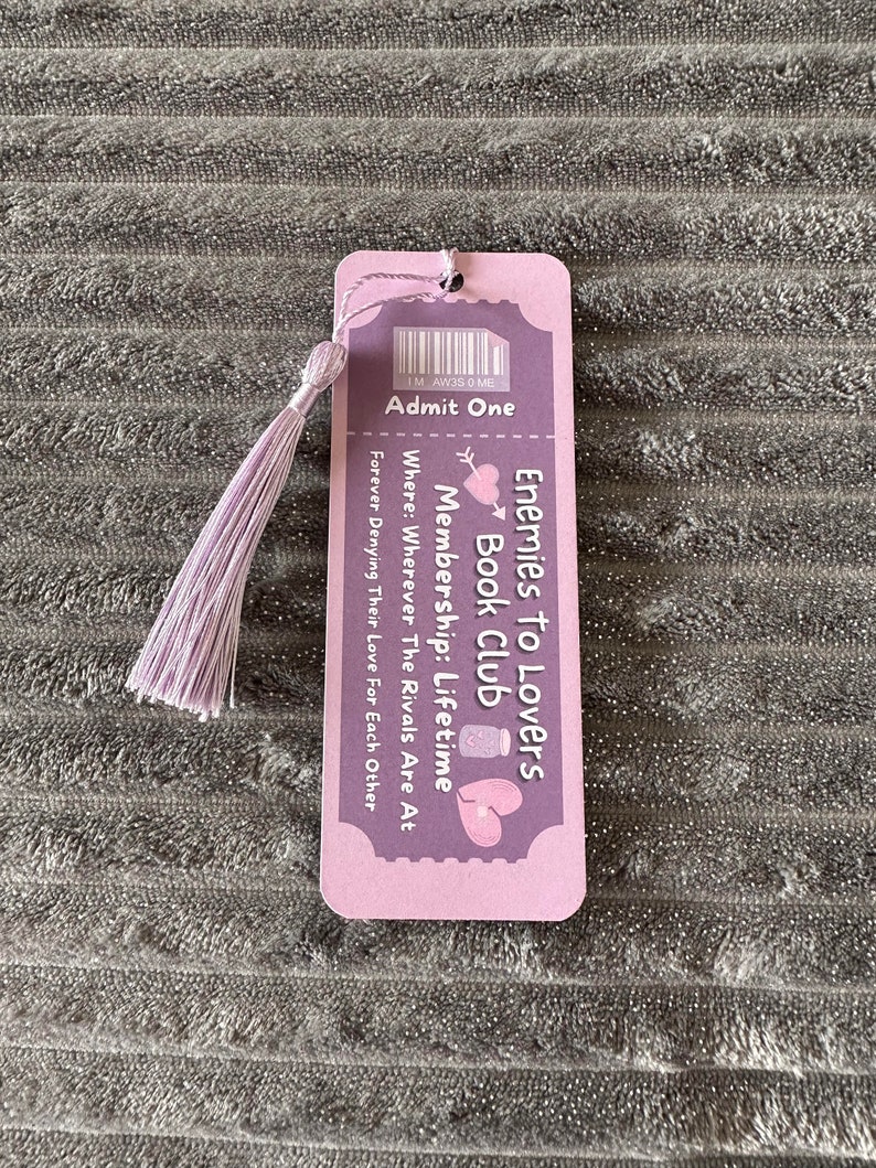 May include: A pink and purple bookmark with a tassel. The bookmark has a ticket design and says "Enemies to Lovers Book Club Membership: Lifetime" and "Where: Wherever The Rivals Are At" and "Forever Denying Their Love For Each Other".
