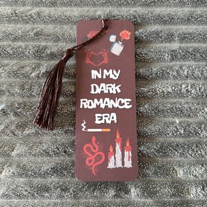 May include: A brown bookmark with a tassel. The bookmark has a dark red background with white text that reads "In My Dark Romance Era". The bookmark is decorated with images of a heart, a lighter, a snake, and candles.