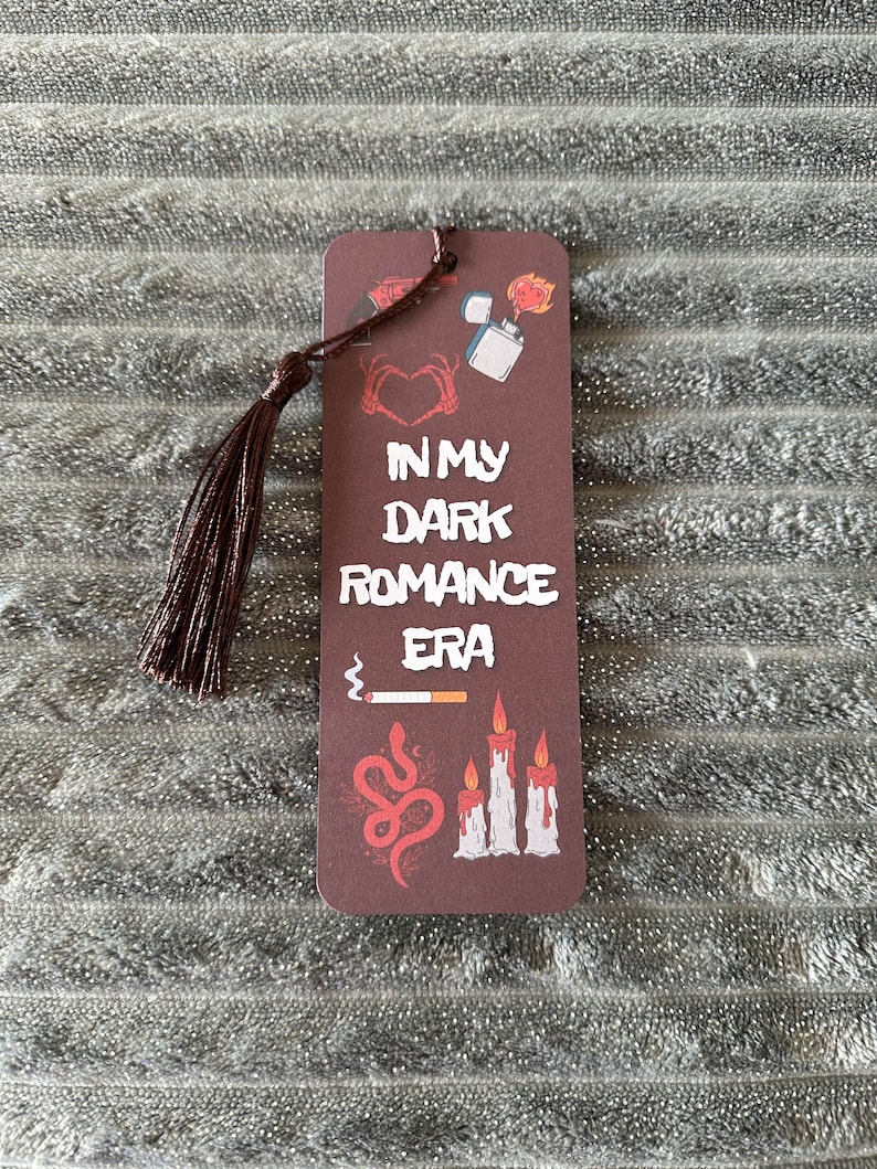May include: A brown bookmark with a tassel. The bookmark has a dark red background with white text that reads "In My Dark Romance Era". The bookmark is decorated with images of a heart, a lighter, a snake, and candles.