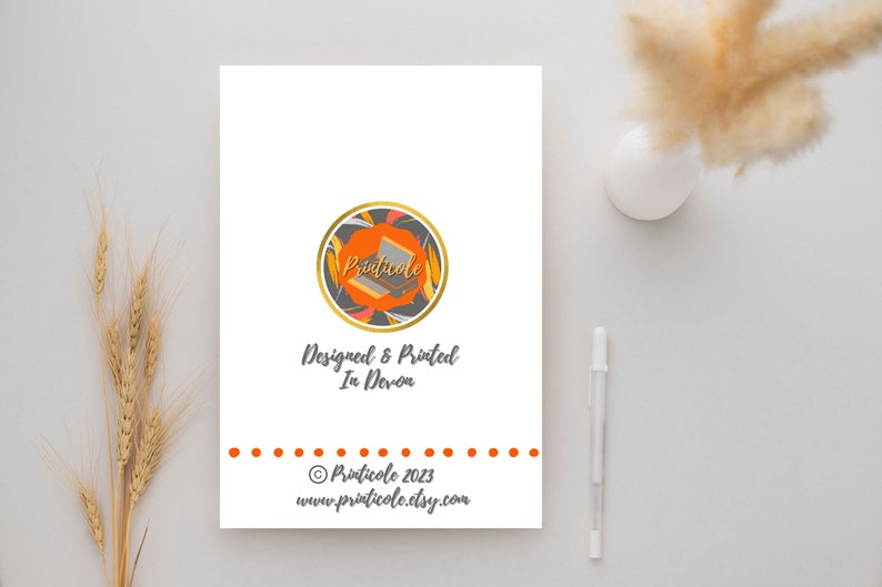 May include: A white card with a logo and text. The logo is a circle with orange, gold, and gray elements. Text includes "Designed & Printed In Devon" and the website address. A pen and decorative plant are in the background.