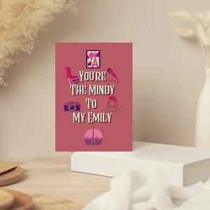 May include: A greeting card with the text "You're the Mindy to my Emily" in white and pink. The card features illustrations of a high-heeled boot, camera, microphone, beret, and the Eiffel Tower. The card is displayed on a white surface.