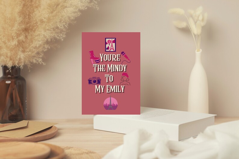 May include: A greeting card with the text "You're the Mindy to my Emily" in white and pink. The card features illustrations of a high-heeled boot, camera, microphone, beret, and the Eiffel Tower. The card is displayed on a white surface.