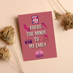 May include: A greeting card with the text "You're the Mindy to my Emily" in white, with illustrations of a high-heeled boot, camera, microphone, and Eiffel Tower. The card is a shade of pink.