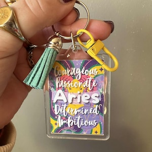 May include: A clear rectangular keychain with the word "Aries" in white, along with the words "Courageous, Passionate, Determined, Ambitious". The keychain has a yellow clasp, a silver keyring, and a mint green tassel.
