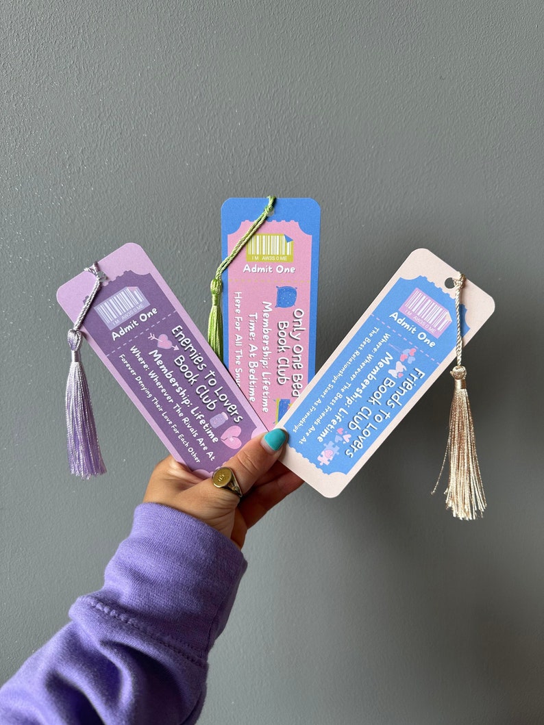 May include: Three colourful bookmarks with a ticket design. Each bookmark has a different book club theme: "Friends to Lovers Book Club", "Enemies to Lovers Book Club", and "Only One Bed Book Club". Each bookmark has a tassel.