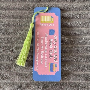 May include: A pink and blue bookmark with a green tassel. The bookmark has a ticket design with the text "Only One Bed Book Club" and "Membership: Lifetime".