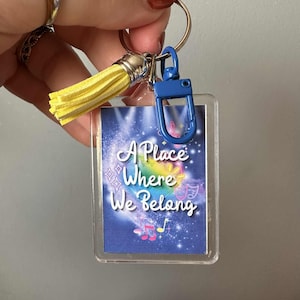 May include: A clear rectangular keychain with a blue clasp and a yellow tassel. The keychain features a colorful design with the text "A Place Where We Belong" in white script, with musical notes and stars.