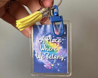 May include: A clear rectangular keychain with a blue clasp and a yellow tassel. The keychain features a colorful design with the text "A Place Where We Belong" in white script, with musical notes and stars.