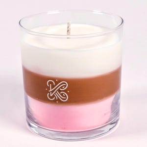 K&ouml;nnte beinhalten: Eine dreischichtige Kerze in einem klaren Glas. Die Schichten sind wei&szlig;, braun und rosa. Ein wei&szlig;es Logo ist auf die braune Schicht gedruckt. Ein Docht ragt aus der Oberseite heraus. Die Kerze steht auf einer hellrosa Oberfl&auml;che.