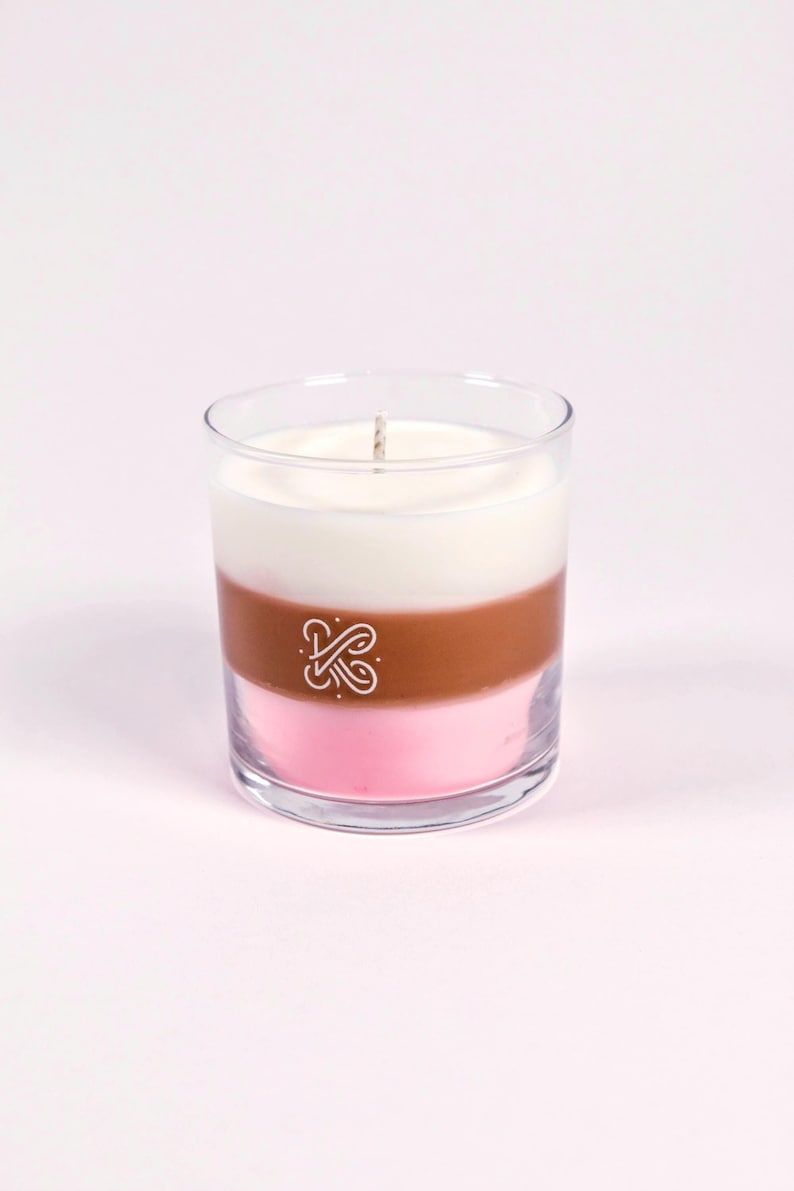 K&ouml;nnte beinhalten: Eine dreischichtige Kerze in einem klaren Glas. Die Schichten sind wei&szlig;, braun und rosa. Ein wei&szlig;es Logo ist auf die braune Schicht gedruckt. Ein Docht ragt aus der Oberseite heraus. Die Kerze steht auf einer hellrosa Oberfl&auml;che.