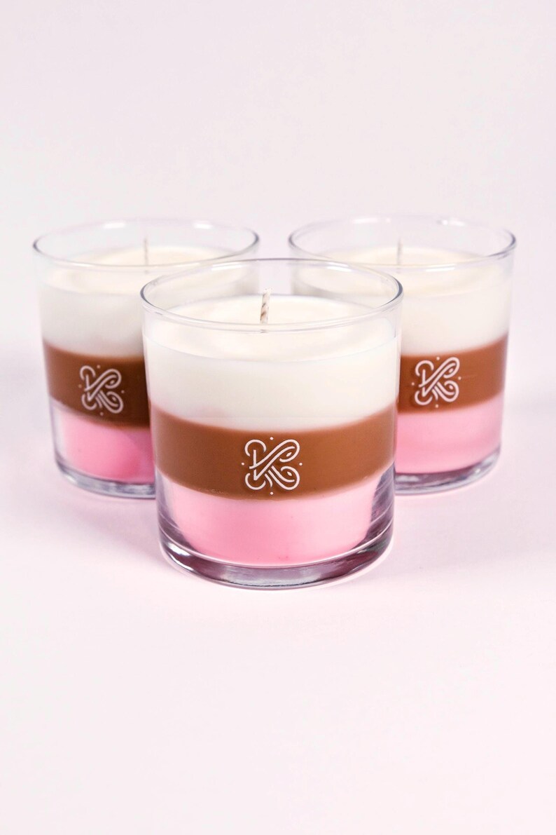 K&ouml;nnte beinhalten: Drei mehrschichtige Kerzen in transparenten Glasbeh&auml;ltern. Die Schichten sind rosa, braun und wei&szlig;. Jede Kerze hat einen wei&szlig;en Docht und ein wei&szlig;es Logo. Wohndekoration.