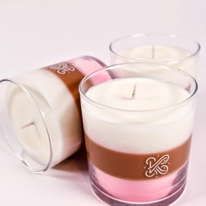 K&ouml;nnte beinhalten: Drei mehrschichtige Kerzen in transparenten Glashaltern. Jede Kerze hat eine wei&szlig;e obere Schicht, eine braune Mittelschicht und eine rosa untere Schicht. Auf der braunen Schicht ist ein Logo aufgedruckt. Die Kerzen haben Dochte.