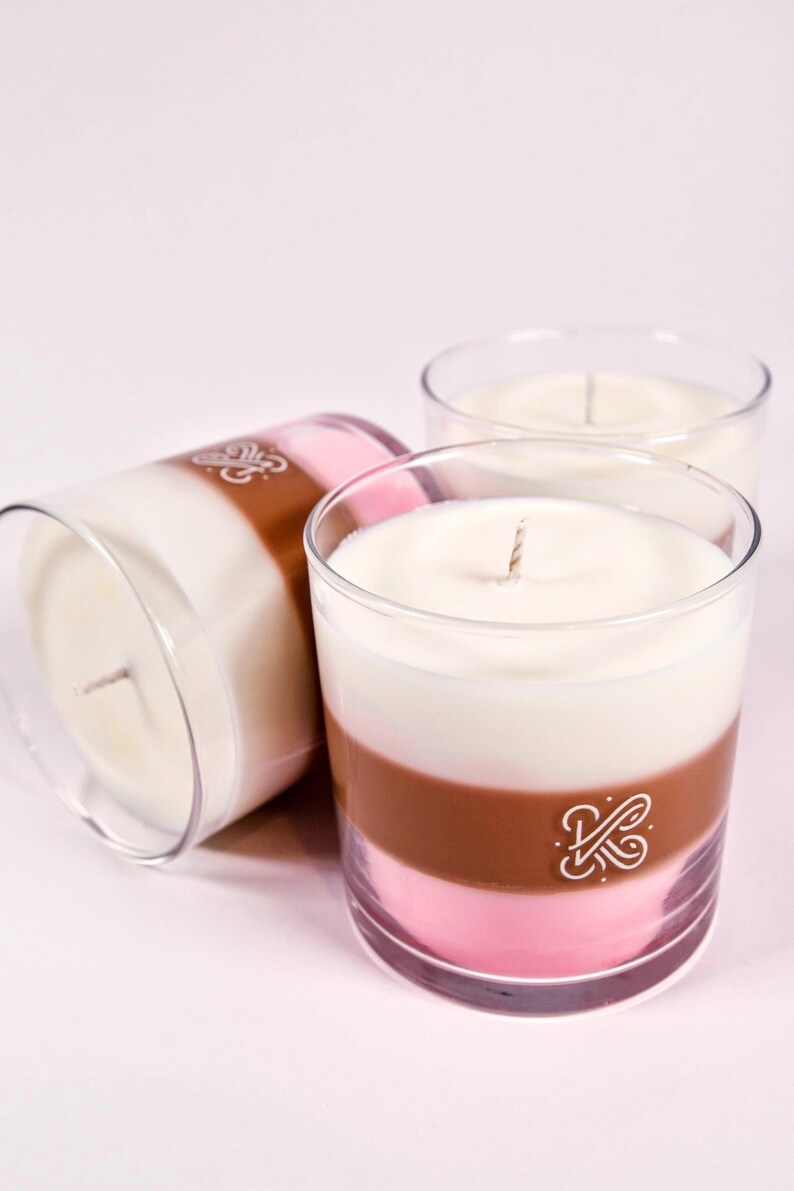 K&ouml;nnte beinhalten: Drei mehrschichtige Kerzen in transparenten Glashaltern. Jede Kerze hat eine wei&szlig;e obere Schicht, eine braune Mittelschicht und eine rosa untere Schicht. Auf der braunen Schicht ist ein Logo aufgedruckt. Die Kerzen haben Dochte.