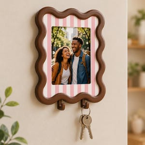 Op de afbeelding: Een bruine fotolijst voor aan de muur met golvende randen en een roze-witte verticaal gestreepte omlijsting. De lijst bevat een foto van een lachend stel en heeft twee haakjes aan de onderkant, waaraan een metalen sleutelbos hangt.