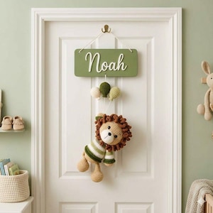 Puede incluir: Un letrero rectangular de madera verde salvia con el nombre Noah en letra blanca, colgado de una cuerda. Debajo del letrero, tres globos pequeños de ganchillo en crema, verde y oliva cuelgan sobre un león de ganchillo con camisa a rayas.