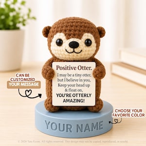 Peut inclure: Une figurine de loutre crochetée marron et crème tenant un panneau carré blanc avec le texte : Positive Otter. I may be a tiny otter, but I believe in you, Keep your head up & float on, YOU'RE OTTERLY AMAZING! Elle repose sur une base bleue.