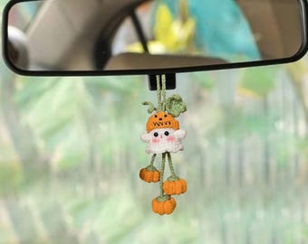 May include: A handmade car charm featuring a white ghost wearing an orange pumpkin hat. The charm has three dangling orange pumpkins and is suspended from a green string. The charm is hanging from a car's rearview mirror.