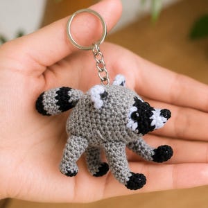 May include: A small, round, crocheted raccoon keychain featuring a gray body, a striped tail, and a black and white face mask. The raccoon has black-tipped paws and a silver-colored metal keyring attached to the top by a short chain.