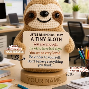 Könnte beinhalten: Eine beige gehäkelte Faultierfigur hält ein rechteckiges Schild mit der Aufschrift LITTLE REMINDERS FROM A TINY SLOTH und fünf positiven Affirmationen in verschiedenen Farben. Das Faultier steht auf einem beigen Sockel mit Gravur YOUR NAME.