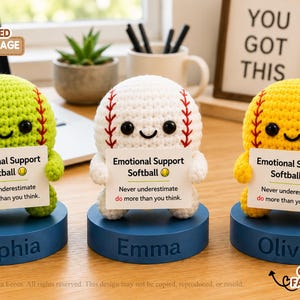 May include: Three crocheted softball figures in lime green, white, and yellow, each with black eyes and a smile. They hold a white sign reading Emotional Support Softball, Never underestimate do more than you think. Each sits on a blue base with names.