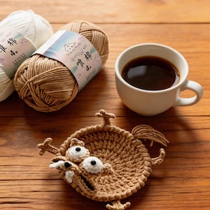 May include: A brown crocheted coaster in the shape of a cartoon character with large white eyes and a wide open mouth. Two balls of yarn and a cup of coffee are also on the wooden surface.