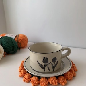 May include: A cream-colored teacup and saucer set with black tulip illustrations. The cup sits on a matching saucer, which rests on an orange crochet coaster. Yarn balls in green, orange, and cream are visible in the background.