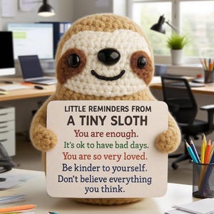 Op de afbeelding: Een gehaakte luiaard figuur, beige en bruin, houdt een bord met inspirerende tekst vast. Het bord luidt "KLEINE HERINNERINGEN VAN EEN KLEINE LUIARD" en bevat positieve affirmaties. De achtergrond is een kantooromgeving.