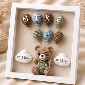 Op de afbeelding: Een witte fotolijst met een gehaakte bruine teddybeer in een groene tuinbroek met vlinderdas. De beer is verbonden met zeven gehaakte ballonnen in bruin, blauw en groen, die de naam MIKE vormen. Twee witte wolken tonen 07.12.2027 en 09:25 PM.