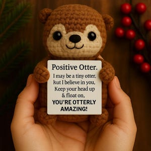 May include: A crocheted brown otter holding a white sign with the text "Positive Otter." The sign reads, "I may be a tiny otter, kut I believe in you, Keep your head up & float on, YOU'RE OTTERLY AMAZING!" The otter has black eyes.
