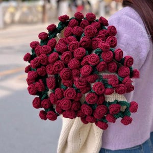 May include: A crocheted tote bag with a design of red roses and green leaves. The bag is being held by a person wearing a purple sweater and blue jeans.
