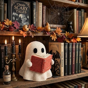 May include: A cream-colored crochet ghost figure with black eyes holding a small orange book. The ghost is positioned on a wooden shelf among various books, including titles like Classic Scary Tales, Frankenstein, and Dracula, with autumn leaves.