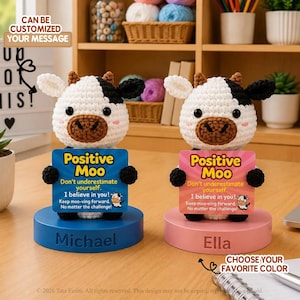 May include: Two crocheted cow figurines with white and black yarn, brown snouts, and black eyes. Each holds a sign reading Positive Moo, Don't underestimate yourself. I believe in you! Keep moo-ving forward. No matter the challenge. Bases are blue and pink.