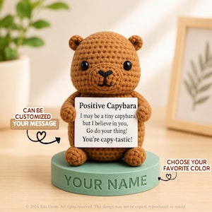 May include: A brown crocheted capybara figurine standing on a round light green base with the text YOUR NAME embossed on the front. The capybara holds a white rectangular sign that reads Positive Capybara, I may be a tiny capybara, but I believe in you, Go do your thing! You're capy-tastic!