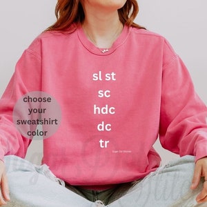 May include: A pink sweatshirt with the text "sl st, sc, hdc, dc, tr" and "Sugar Dot Stitches" printed on the front. The text "choose your sweatshirt color" is in a circle on the left side of the sweatshirt.