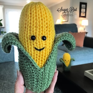 May include: A knitted corn on the cob plush toy with a smiling face. The corn is yellow and the husk is green. The toy is being held by a hand.