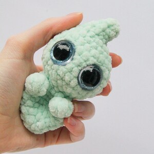 Puede incluir: Un peque&ntilde;o juguete de peluche tejido a crochet de color verde claro con grandes ojos azules brillantes. El juguete tiene la forma de una criatura con una cabeza larga y puntiaguda y un cuerpo peque&ntilde;o.