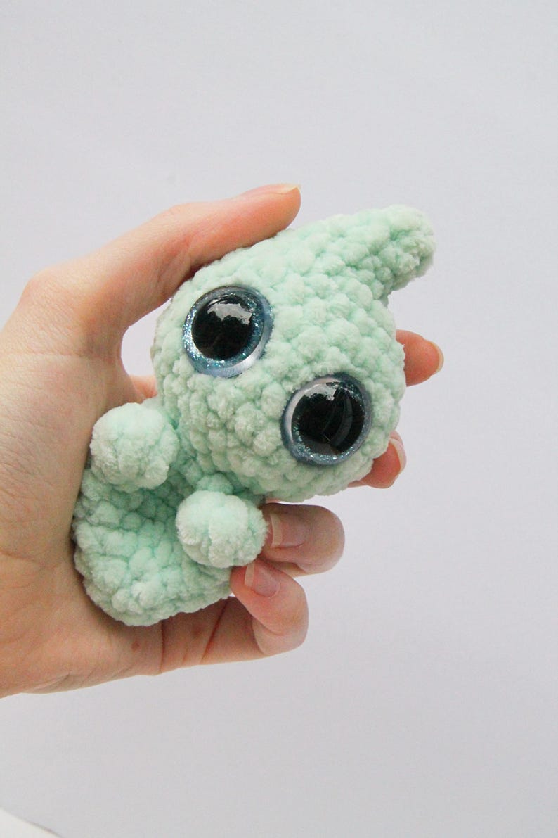 Puede incluir: Un peque&ntilde;o juguete de peluche tejido a crochet de color verde claro con grandes ojos azules brillantes. El juguete tiene la forma de una criatura con una cabeza larga y puntiaguda y un cuerpo peque&ntilde;o.
