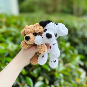 以下が含まれることがあります： 柔らかくふわふわした毛糸で作られた2匹の小さな犬のぬいぐるみ。1匹は黒い鼻と暗い目をした無地のライトブラウンの犬で、もう1匹は黒い斑点、黒い耳のパッチ、黒い鼻を持つ白い犬です。どちらも垂れ耳と丸い目をしています。