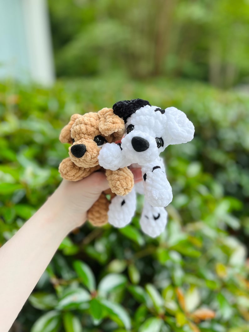 以下が含まれることがあります： 柔らかくふわふわした毛糸で作られた2匹の小さな犬のぬいぐるみ。1匹は黒い鼻と暗い目をした無地のライトブラウンの犬で、もう1匹は黒い斑点、黒い耳のパッチ、黒い鼻を持つ白い犬です。どちらも垂れ耳と丸い目をしています。
