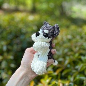 May include: A handmade crochet dog toy in black, white, and gray. The small stuffed animal has a fluffy white body and paws, with a black and gray head. The dog is held in a person's hand against a blurred green background.