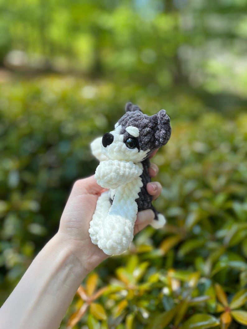 May include: A handmade crochet dog toy in black, white, and gray. The small stuffed animal has a fluffy white body and paws, with a black and gray head. The dog is held in a person's hand against a blurred green background.