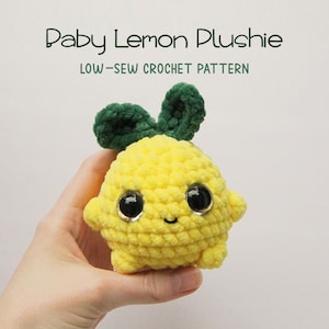 May include: A crocheted lemon plush toy with a green leaf, black eyes, and a smiling mouth. The toy is being held in a hand. The text "Baby Lemon Plushie" and "LOW-SEW CROCHET PATTERN" is above the toy.