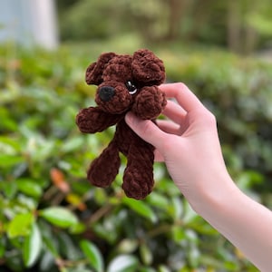 以下が含まれることがあります： 柔らかい質感の糸で作られた、小さなダークブラウンの犬のぬいぐるみ。このぬいぐるみは、垂れ耳、黒い鼻を持つ丸みを帯びた鼻先、そして小さく暗い光沢のある目を特徴としています。かぎ針編みの構造により、ループ状の模様がはっきりと見えます。