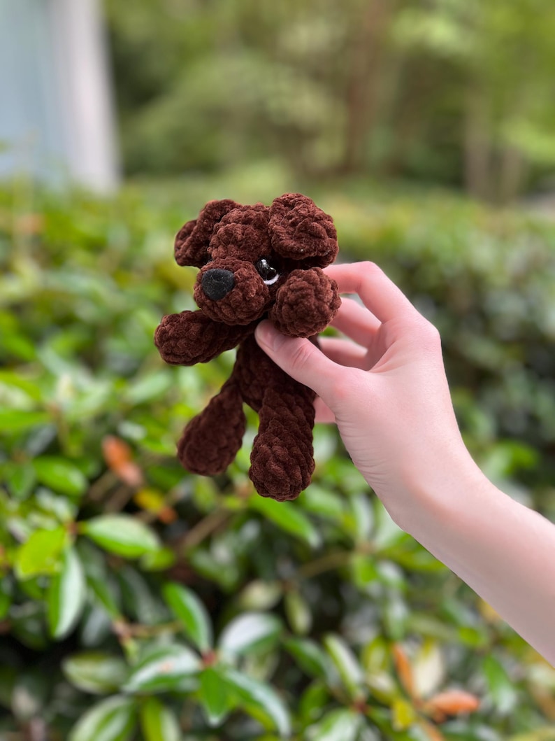 以下が含まれることがあります： 柔らかい質感の糸で作られた、小さなダークブラウンの犬のぬいぐるみ。このぬいぐるみは、垂れ耳、黒い鼻を持つ丸みを帯びた鼻先、そして小さく暗い光沢のある目を特徴としています。かぎ針編みの構造により、ループ状の模様がはっきりと見えます。