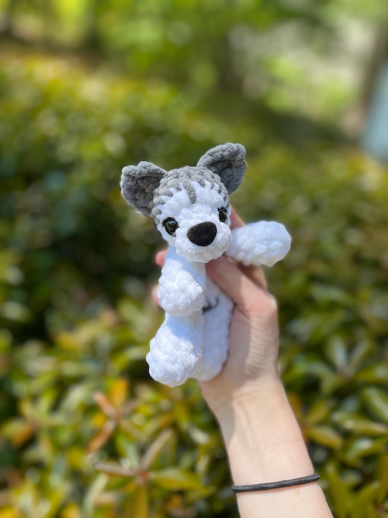 May include: A handmade crochet dog toy in white and gray. The toy features a black nose, small black eyes, and gray ears. The dog is in a sitting position with its paws extended. The background is a blurred green.
