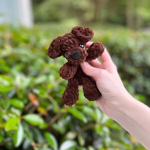 以下が含まれることがあります： 柔らかい質感の糸で作られた、ダークブラウンの小さな犬のぬいぐるみ。このぬいぐるみは、垂れ下がった耳、黒い鼻を持つ丸みを帯びた鼻先、そして小さな暗い色の目が特徴です。手足がはっきりと見えるシンプルなデザインのぬいぐるみです。