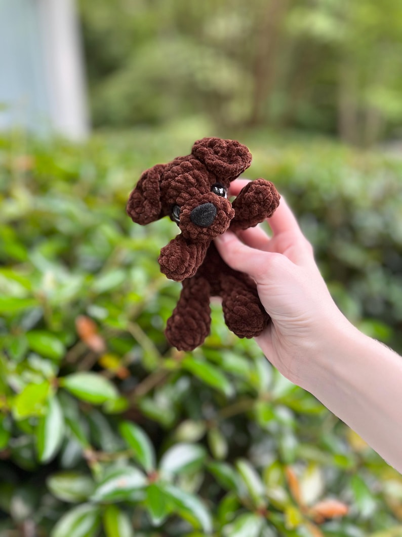 以下が含まれることがあります： 柔らかい質感の糸で作られた、ダークブラウンの小さな犬のぬいぐるみ。このぬいぐるみは、垂れ下がった耳、黒い鼻を持つ丸みを帯びた鼻先、そして小さな暗い色の目が特徴です。手足がはっきりと見えるシンプルなデザインのぬいぐるみです。