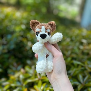 May include: A handmade crochet dog toy in white, brown, and black. The dog has a white body, brown ears and head markings, and black eyes and nose. The toy is held up against a blurred green background.