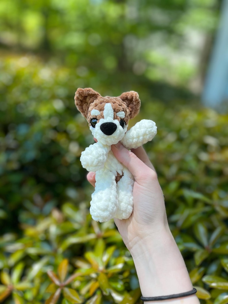 May include: A handmade crochet dog toy in white, brown, and black. The dog has a white body, brown ears and head markings, and black eyes and nose. The toy is held up against a blurred green background.
